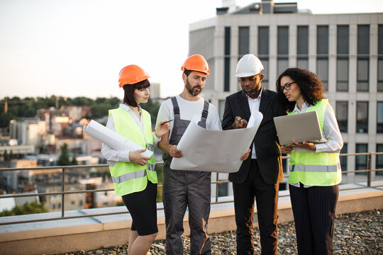 Group of four multiracial people in suits and helmets brainstorming for common project of new real estate building. Construction team working with blueprints and modern laptop in fresh air.