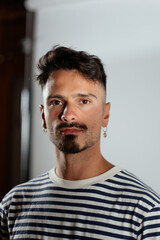 Fototapeta premium Portrait of a man in a studio. Behind him is a white backdrop. He is dressed casually, with earrings and a striped shirt. He has a goatee and a fade haircut. He looks relaxed, with a hint of a smile.