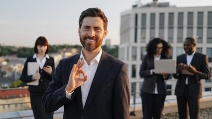 Portrait of confident bearded executive in business attire posing on camera smiling, showing sign ok. International colleagues of mixed nationality standing behind and operating modern gadgets.