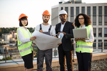 Portrait of multicultural group of developers posing on camera with portable gadget and blueprints. Four international colleagues in vests and hardhats smiling while meeting at panoramic terrace.
