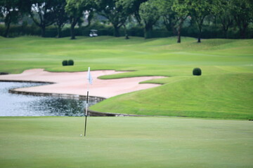 A golf ball that almost hits the hole with the flag. On the green with many signs of repair In the background are sand bunkers, ponds and lush lawns. There is a blurred golf cart in the distance.