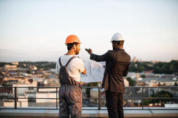 Back view of foreman in grey uniform and business investor in black suit watching together at engineering drawings while standing on panoramic terrace. Male builders discussing real estate project.
