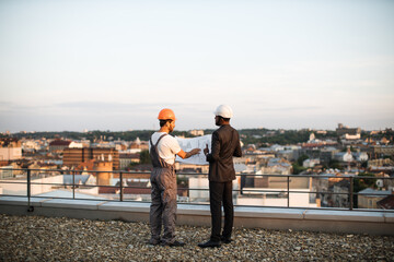 Back view of foreman in grey uniform and business investor in black suit watching together at engineering drawings while standing on panoramic terrace. Male builders discussing real estate project.