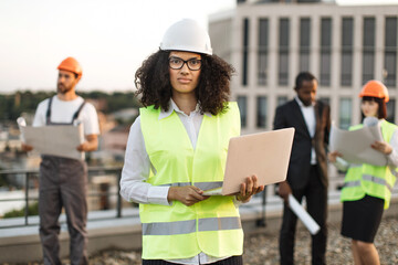 Portrait of african american female urban planner in protective wear and glasses holding remote computer and looking at camera. Diverse architects reviewing building blueprints on roof terrace.