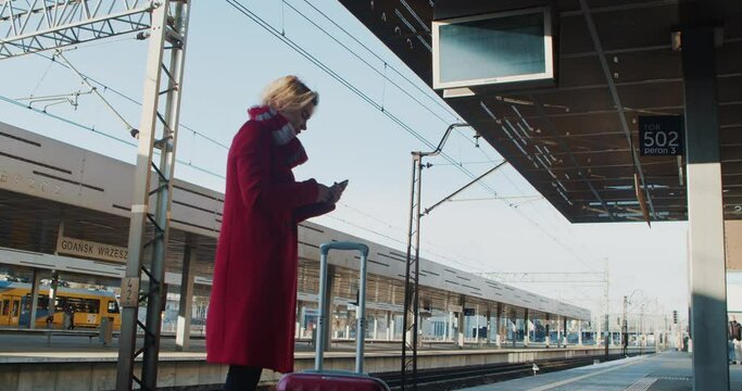Young Woman in a Red Coat with Luggage is a Passenger Checking the Timetable at the Railway Stop. Young Woman in a Red Coat the Stop of the Railway Station of Public Transport Looks at the Schedule. 
