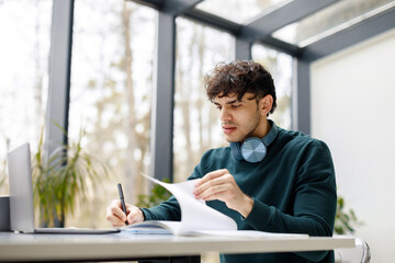 Student guy writing homework assignment in copybook, watching webinars online on laptop, studying in modern library