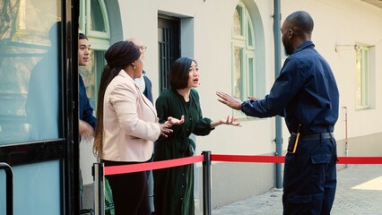 Security guard asking people to wait, trying to reason with crowd of anxious clients waiting since...