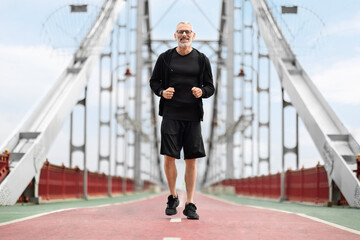 Happy well-fit senior sportsman jogging by city bridge, copy space