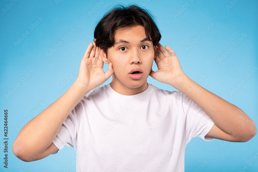 Wall mural asian teen guy listening holding hands near ears, blue background