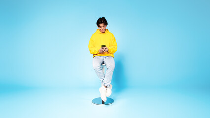 Fototapeta na wymiar Chinese Teen Guy Using Smartphone Websurfing Sitting On Blue Background