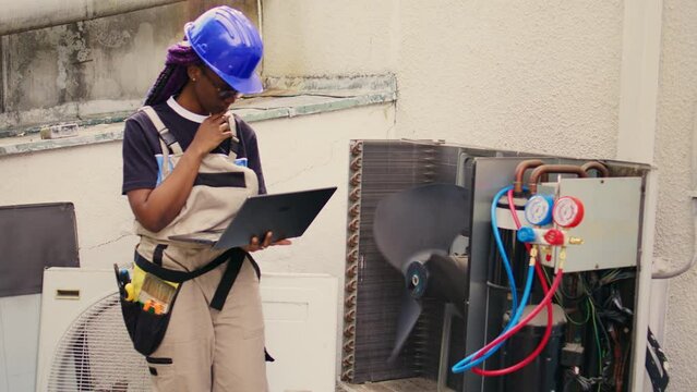 Qualified african american electrician working on outside hvac system, holding laptop. Licensed serviceman optimizing external air conditioner performance, ensuring it operates at maximum capacity