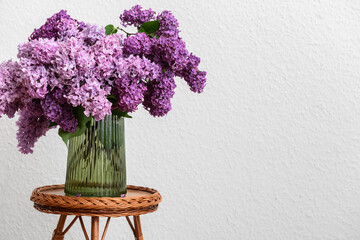Vase with beautiful lilac flowers on table near white wall in room, closeup