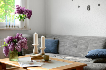 Interior of light living room with beautiful lilac flowers, candles, table and sofa