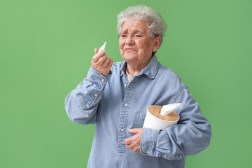 Ill senior woman with nasal drops and tissue box on green background