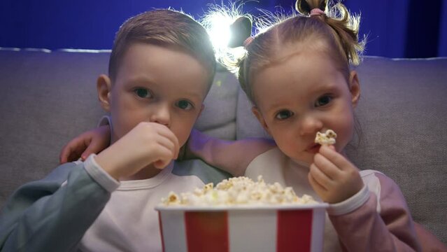 Portrait of cute brother sister watches video on movie projector eats popcorn.