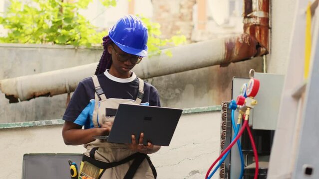 Expert repairman servicing external HVAC system internal components, checking freon level and calibrating thermostats. Proficient worker imputing maintenance results data on laptop