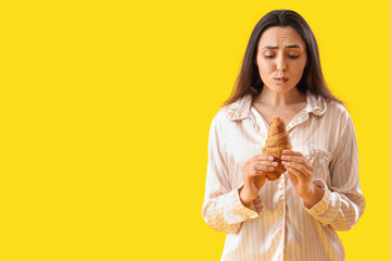 Worried young woman in pajamas with tasty croissant on yellow background