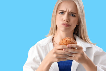 Worried young woman with tasty muffin on blue background, closeup