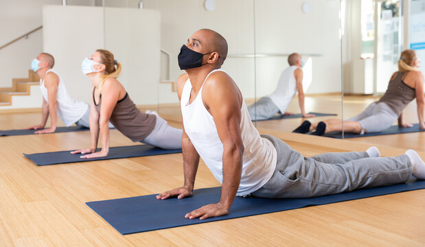 Hispanic man wearing protective mask performing yoga exercises during group workout in gym. Healthy lifestyle and pandemic precautions concept..
