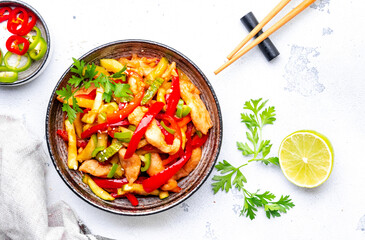 Asian cuisine stir fried chicken breast, red paprika and zucchini with sesame seeds, lime and soy sauce in ceramic bowl. White table background, top view