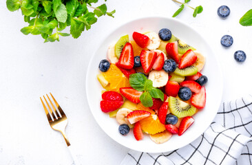 Fruit and berry salad with fresh strawberries, blueberries, banana, kiwi, orange and mint leaves, white table background, top view