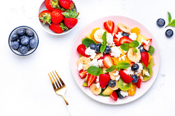 Summer fruit and berry salad with fresh strawberries, blueberries, banana, cottage cheese and mint on white table background, top view
