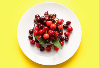 Plate and glass bowl with sweet cherries on yellow background