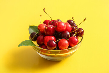 Glass bowl with sweet cherries and leaves on yellow background