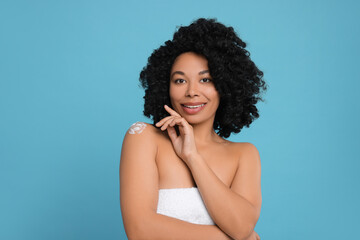 Young woman applying body cream onto shoulder on light blue background