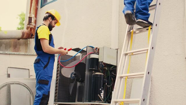 Proficient expert holding electric power drill stepping down from folding ladder after finishing servicing rooftop hvac system coworker assembles manifold gauges to check freon levels.