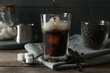 Pouring coffee into glass with ice cubes, mint and sugar on wooden table