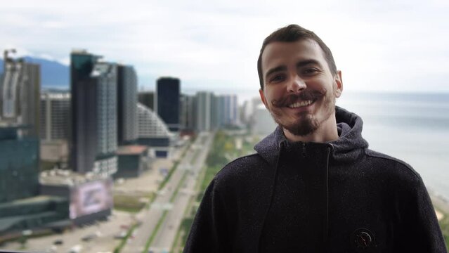 Portrait of caucasian man standing on balcony against coastal city background. Young adult male person with moustache in warm sweater is looking at camera and smiling. Visit Georgia and stay at hotel.
