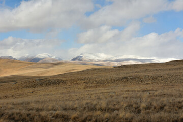 Fototapeta premium Gentle slopes of a hilly steppe with yellowed grass at the foot of snow-capped mountain peaks on a sunny autumn day.