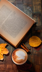 Naklejka na ściany i meble A cup of latte with design sprinkled with cocoa, next to an old book on a wooden table and autumn leaves