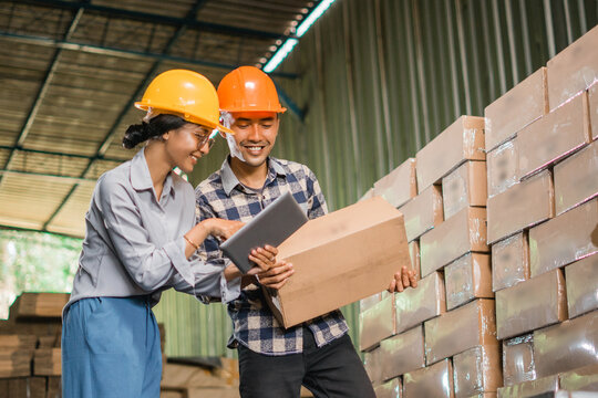two factory workers wear safety helmets while using a tablet and carrying cardboard boxes in a warehouse storage of goods