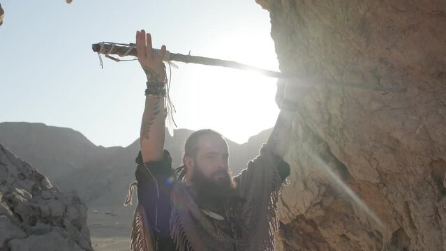 A young bearded man with a staff in his hands in the clothes of a shaman prays on a rock among the sandy dunes of the desert