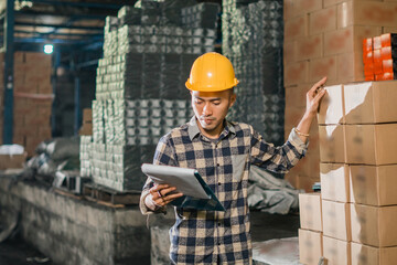 Asian man factory employee wearing helmet working to check data with goods in storage warehouse