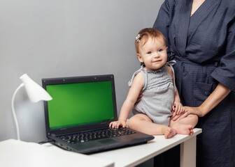 Little gray-eyed 9-month-old girl sitting next to a laptop with a green screen. Chroma key. The...