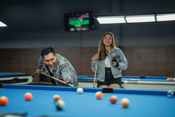 female pool player holding the glass of drink while standing behind the male pool player that poking the white ball