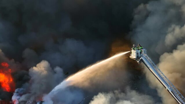 Cinematic drone shot of two firefighters on ladder spraying water on burning building while dark toxic fumes rising up - Aerial top down zoom shot