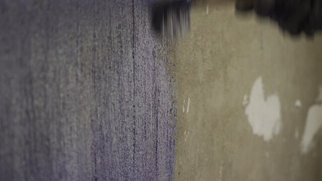 The man's hand is in close-up priming the concrete wall with an old brush in front of the plaster