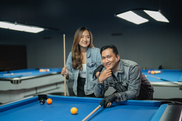 Fototapeta premium female pool player holding the cue stick while standing beside the male player that leaning on the pool