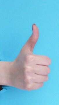 Vertical video. Approval like. Well done. Super excellent. Female hand showing thumb up in breakthrough paper hole torn wallpaper on blue background with free space loop.