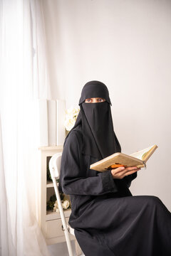 muslim woman wearing a black hijab or niqab and long black dress sitting in a chair and reading book or al qur'an looking at side