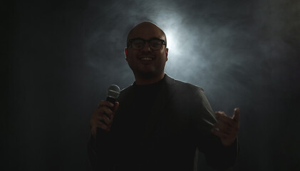 Confident successful speaker man talking on stage with spotlight strike through the darkness at corporate business event. Public speaker giving talk at conference hall. Stand up comedian.