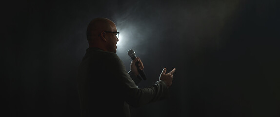Confident successful speaker man talking on stage with spotlight strike through the darkness at corporate business event. Public speaker giving talk at conference hall. Stand up comedian. - Powered by Adobe