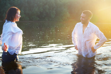 A beautiful adult couple has fun in nature in the water in a river or lake in the summer evening at sunset. A guy and a girl swim and relax outdoors in clothes in white shirts and jeans