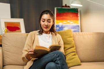 Asian beautiful woman sitting reading book on cozey couch sofa in living room with warm light at night. People female stay home part time relax at home.