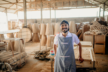 smiling Asian male entrepreneur standing wearing an apron in a woodcraft warehouse