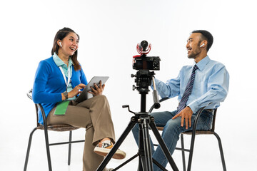 a female journalist in blue cardigan and an adult man in blue shirt sitting on chair while doing an...